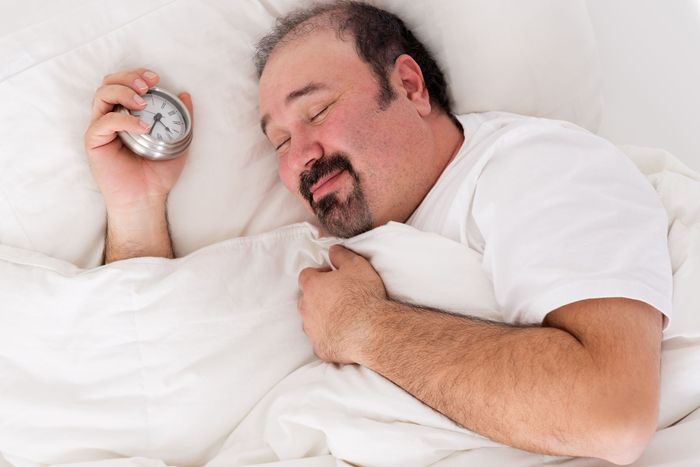 Man lying in bed smiling in contentment after a good nights sleep feeling relaxed and refreshed as he struggles to wake up in the morning with his alarm clock in his hand